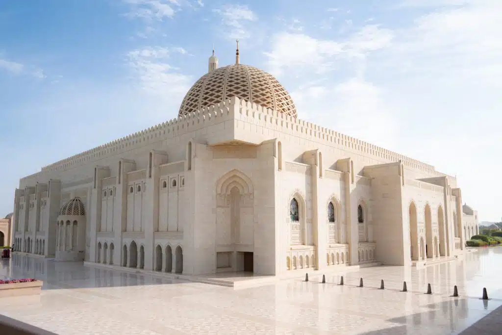 A large, ornate mosque with a grand dome and arched windows stands majestically, crafted from light-colored stone. Its polished courtyard reflects the clear blue sky, making it a must-see for any 1-week Oman itinerary.