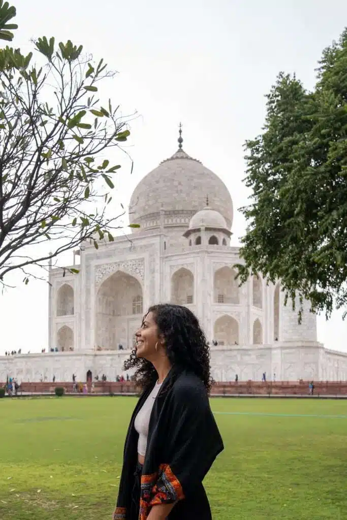 A person with curly hair and a dark shawl stands smiling in front of the Taj Mahal. The iconic white marble mausoleum is surrounded by green trees and a grassy area. The sky is overcast.
