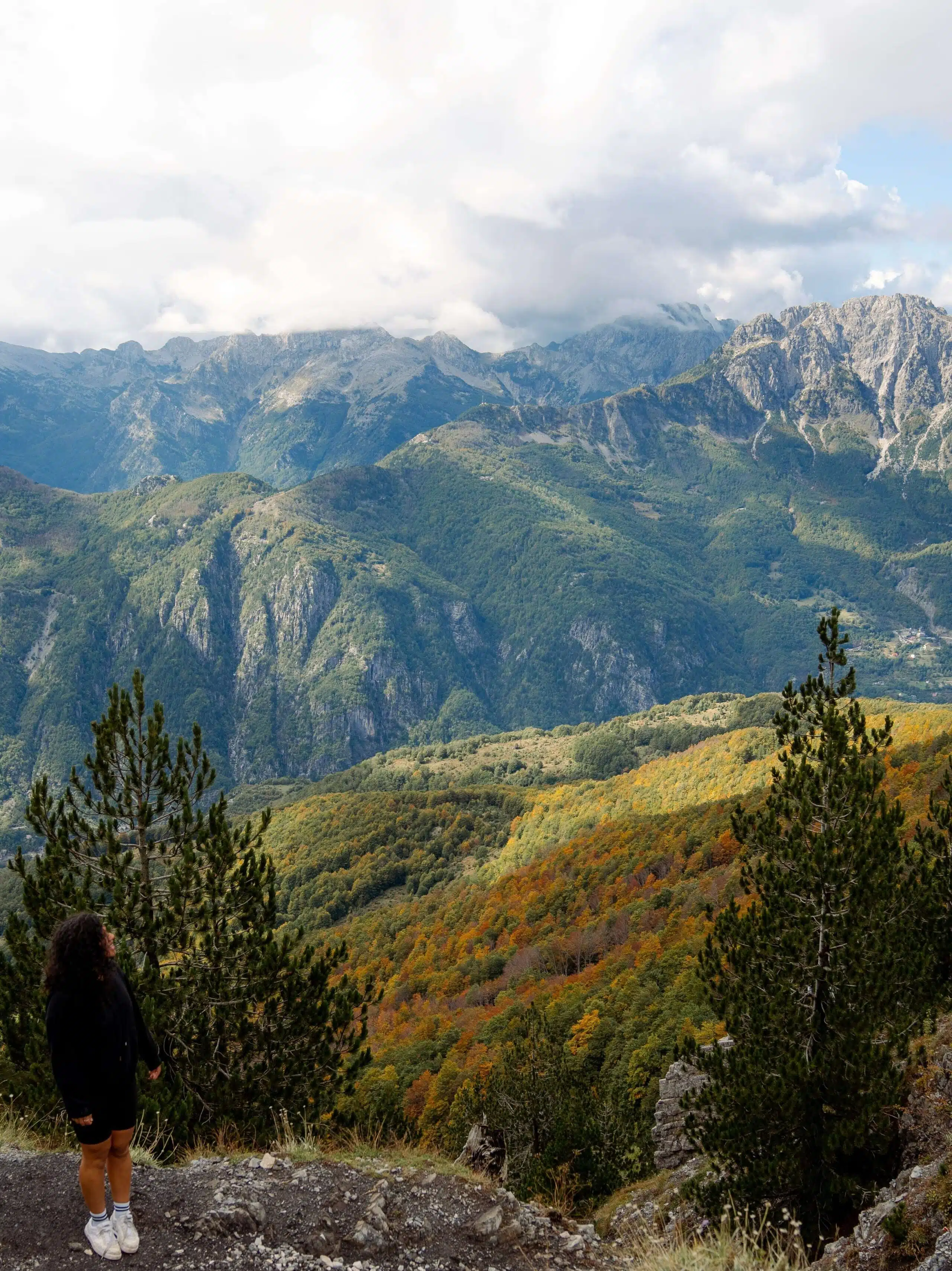 A person stands on a rocky outcrop, gazing at the vast mountainous landscape of their 10-Day Albania Itinerary. The scene features lush green and autumn-colored trees, steep rocky cliffs, and a partly cloudy sky, creating a tranquil and majestic view. Valbona to Theth hike in Albania