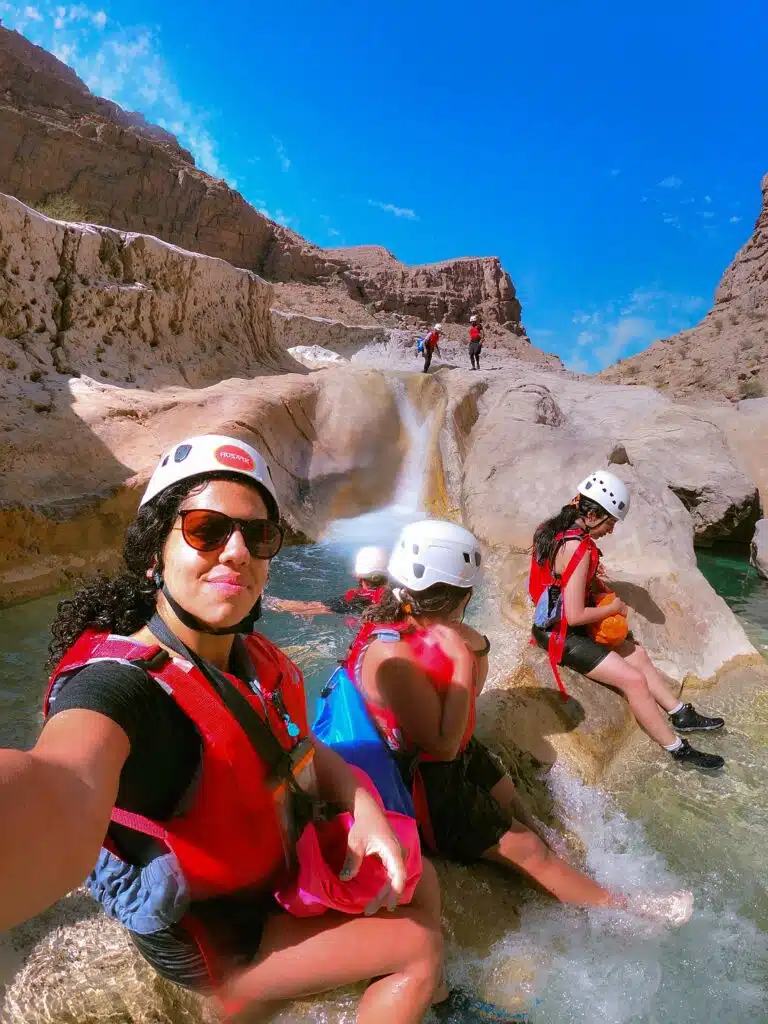 A group of people wearing helmets and life jackets relax on rocks beside a small waterfall in a rocky canyon, part of their 1-week Oman itinerary. The sky is bright blue, and the atmosphere is sunny and adventurous. Part of a trip with Husaak Adventures