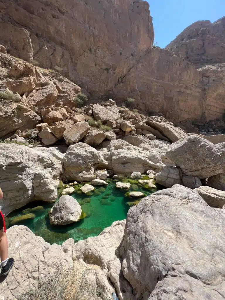 A scenic view of a rocky canyon in Oman, part of a 1-week itinerary, showcases clear turquoise water pools surrounded by large, rugged stones and towering rock formations. The sky is clear and blue above, with part of a person's leg visible on the left edge of the image - Part of a trip with Husaak Adventures