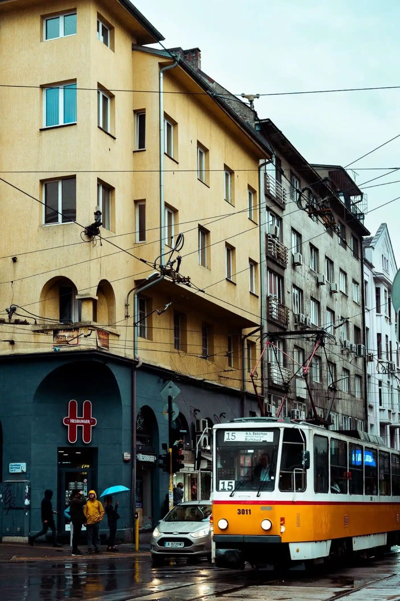One Day In Sofia, a yellow tram glides through a rain-soaked city street lined with buildings. Pedestrians stroll under umbrellas while a few cars are parked nearby. The overcast sky mirrors the tram and architecture in the shimmering wet road.