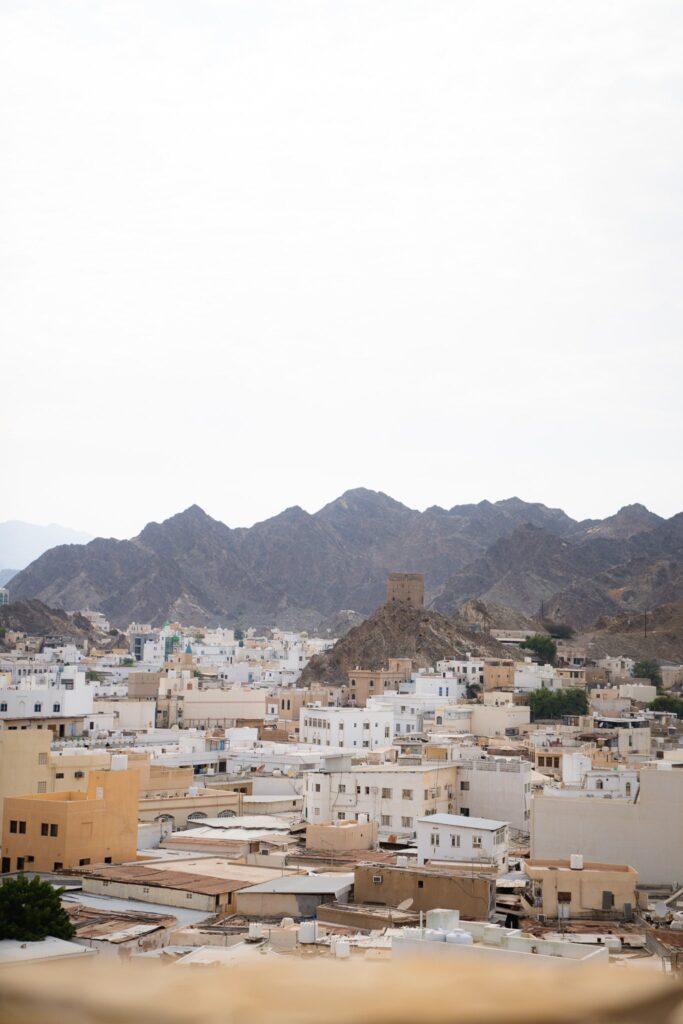 A cityscape with flat-roofed white and beige buildings, a historic stone tower on a hill, and rugged mountains in the background under a cloudy sky—an ideal view to inspire your 1-week Oman itinerary.
