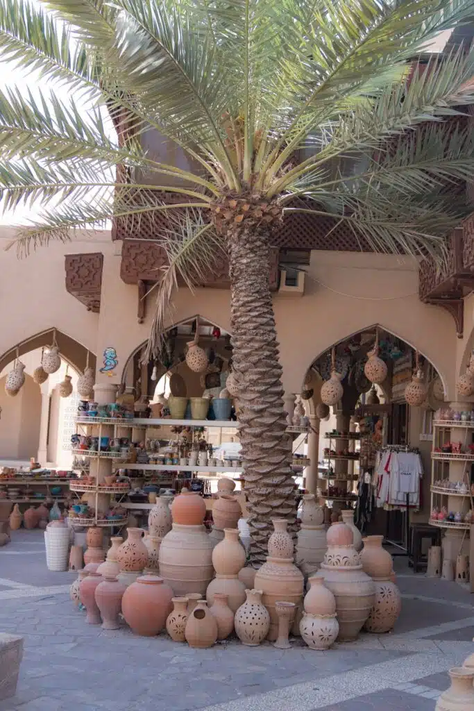 A tall palm tree stands in front of a shop displaying various clay pots, vases, and lanterns—an inviting stop for anyone exploring a 1-week Oman itinerary, with arched doorways and traditional Middle Eastern architectural details.