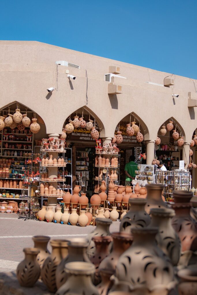 A market stall displays a variety of clay pots and traditional pottery under arched beige buildings, offering an authentic glimpse into local crafts—an ideal stop on your 1-week Oman itinerary beneath a clear blue sky.