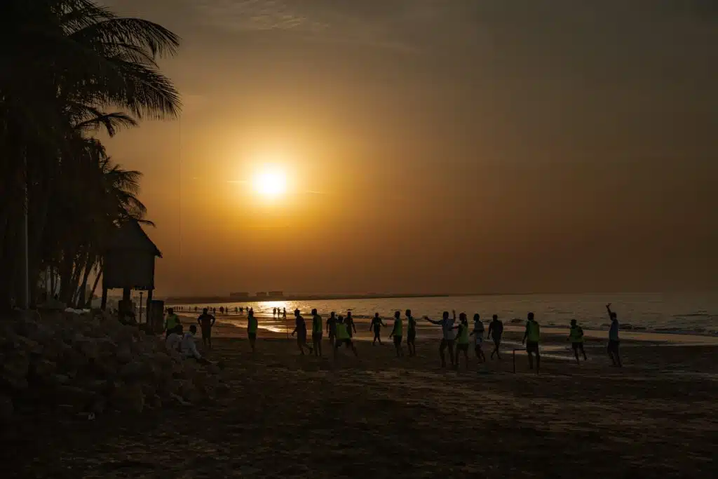A group of people play a game on a sandy beach at sunset, with the sun low in the sky, palm trees to the left, and gentle waves in the background—an ideal moment from a 1-week Oman itinerary.