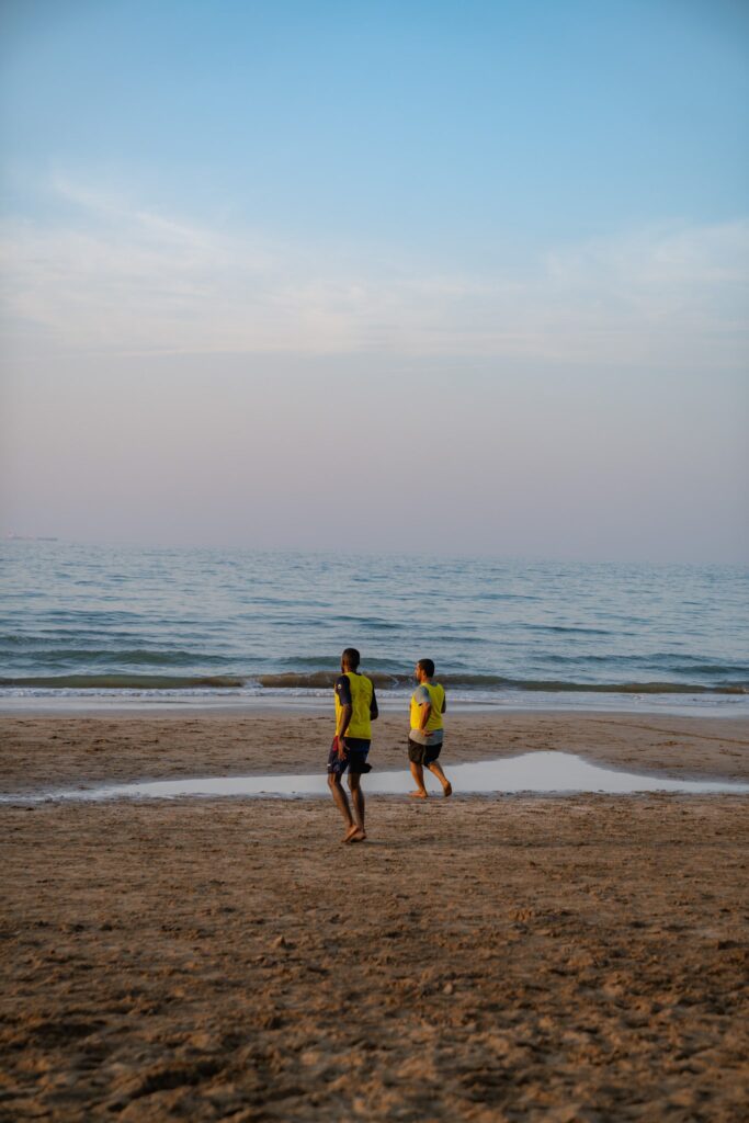Two people in yellow shirts walk barefoot along the sandy beach near the water’s edge, enjoying gentle waves and a clear blue sky—a perfect scene from a 1-week Oman itinerary.