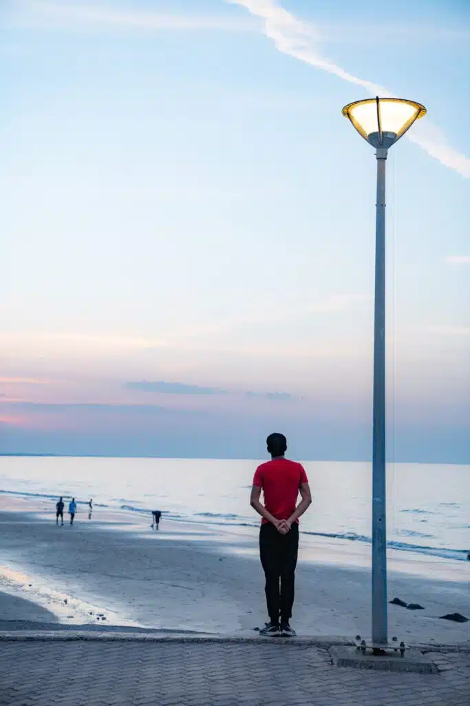 A person in a red shirt stands near a lit streetlamp, gazing at the ocean at sunset—perhaps reflecting on their 1-week Oman itinerary—as a few people stroll along the pastel-colored beach in the background.
