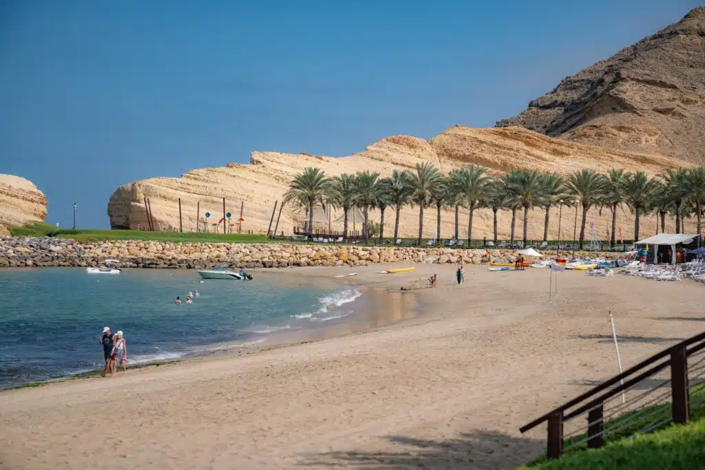 A sandy beach with people walking and swimming, lined with palm trees and backed by rocky hills; boats and beach equipment dot the shore under a clear blue sky, making it an ideal stop on your 1-week Oman itinerary.