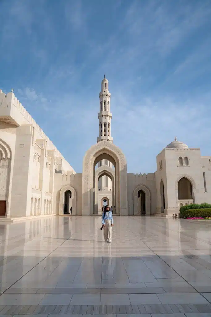 A person walks across a wide marble courtyard in front of a grand mosque with a tall minaret and ornate arches under a bright blue sky, capturing the essence of any 1-week Oman itinerary.