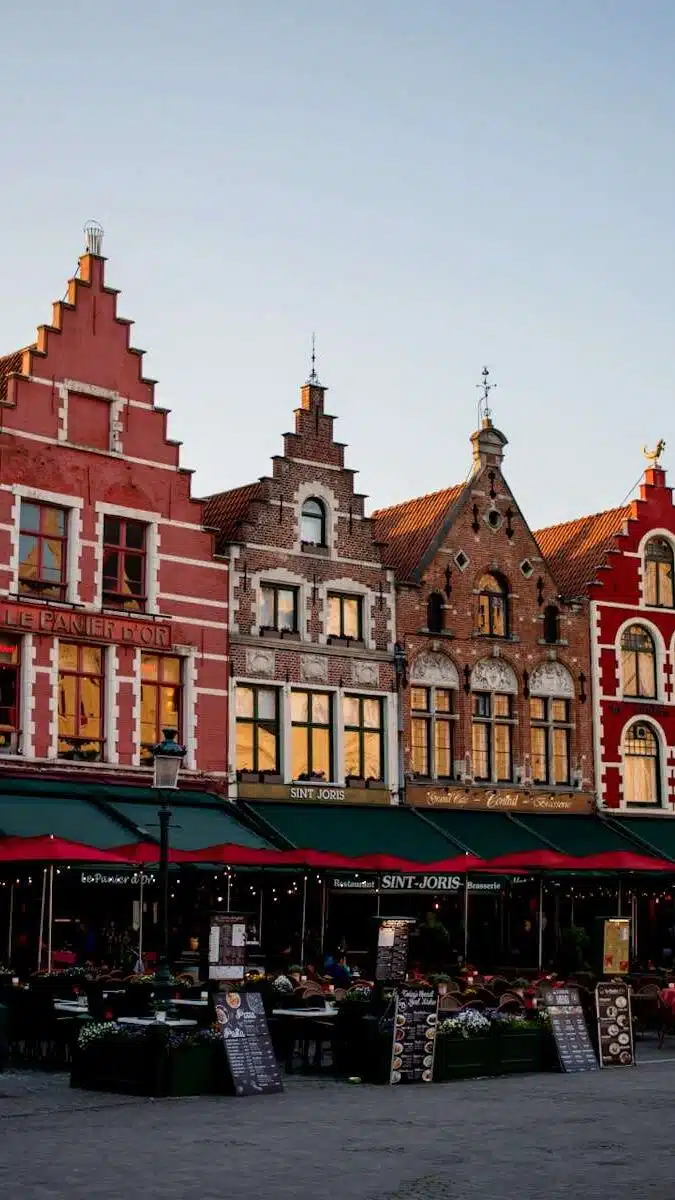 A row of colorful, historic gabled buildings in a located in Markt Square Bruges. The structures feature stepped facades and house cafes with outdoor seating. The sky is clear, adding a warm glow to the scene.