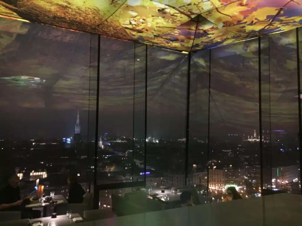 A dimly lit restaurant with large glass windows showcases a panoramic city skyline at night, reminiscent of a stop on your 1-week Eastern Europe itinerary. The ceiling displays a vivid autumn leaf pattern as diners enjoy the view of illuminated buildings and a distant spire.