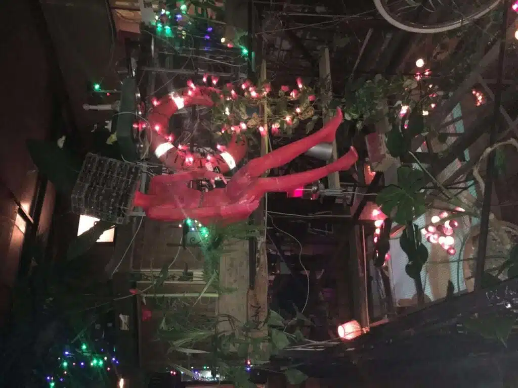 A dimly lit space adorned with colorful string lights and eclectic decorations like a red mannequin, greenery, and even hints of a 1-week Eastern Europe itinerary in the form of travel memorabilia. The artistic ambiance includes a life ring and bicycle wheel, inviting exploration.