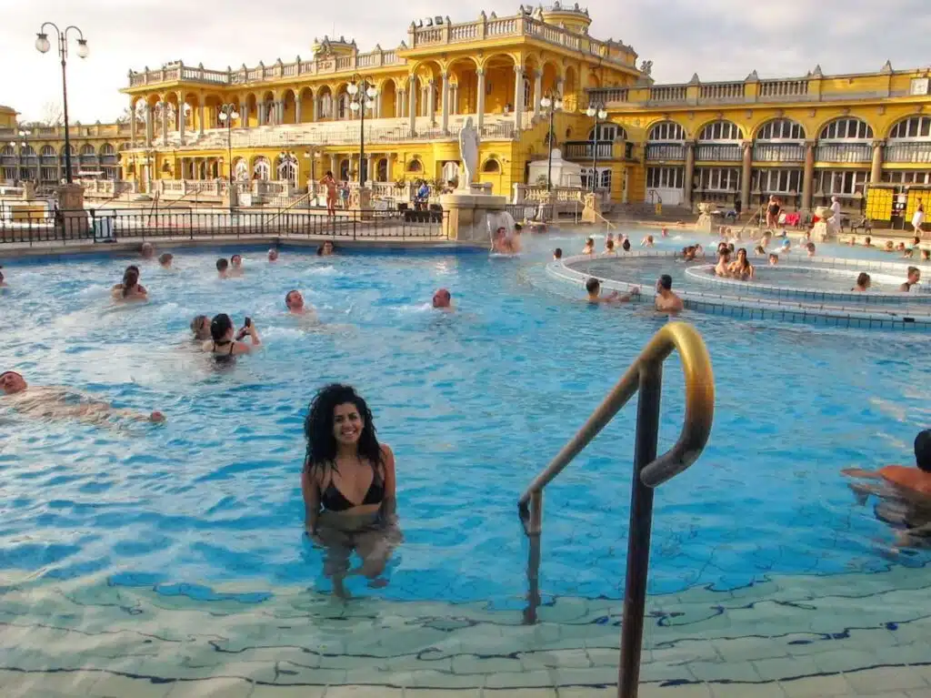 A woman in a swimsuit smiles while enjoying a large, ornate outdoor pool, part of her 1-week Eastern Europe itinerary. The historic yellow building in the background adds charm as others swim and relax. Steps elegantly lead into the inviting water, completing this scenic stop on her adventure.