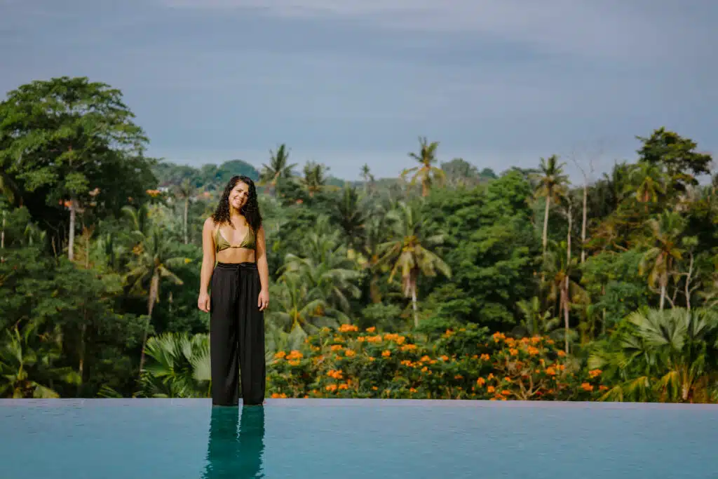 A woman in a bikini top and black pants stands at the edge of an infinity pool, surrounded by lush green trees and tropical plants under a blue sky.