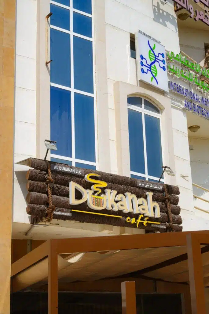 A building facade in Muscat featuring a vertical window and two business signs: "Dukanah Café" with a coffee cup logo, and an international health and biotechnology center—perfect stops during 24 hours in Muscat.
