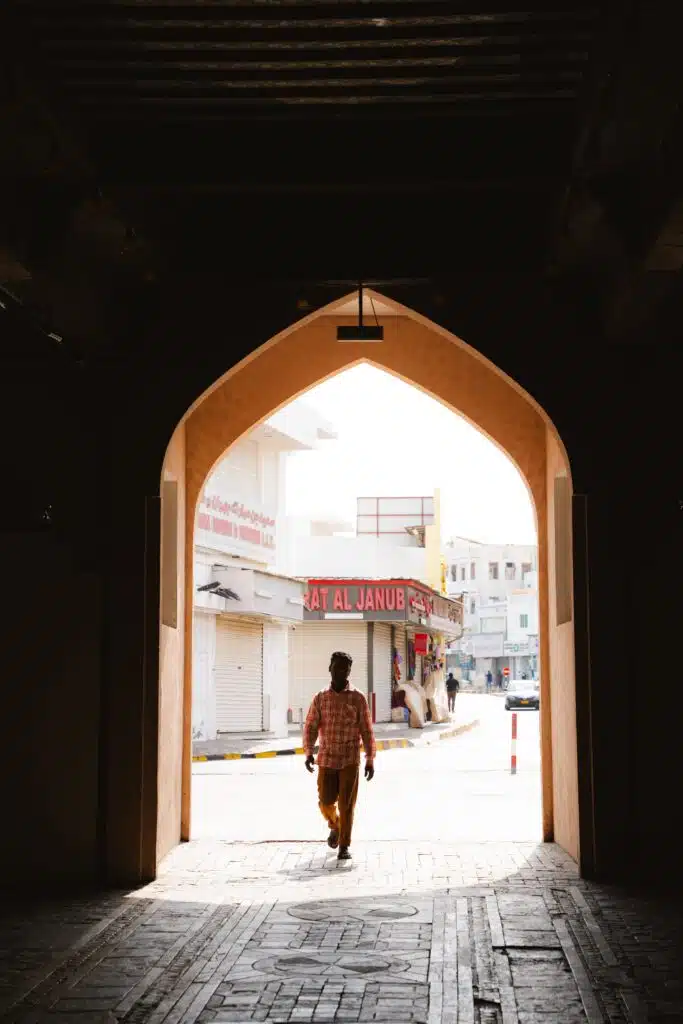 A person walks through an arched doorway into a sunlit street lined with shops, capturing the vibrant atmosphere of spending 24 hours in Muscat, with bright sunlight contrasting sharply against the shadowed archway.