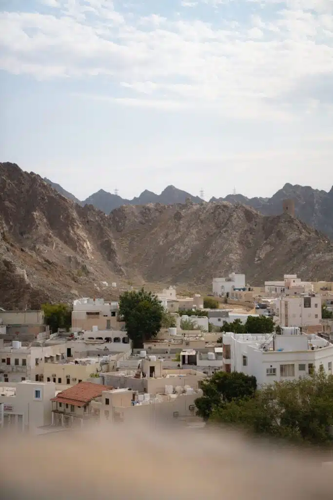 A town with white buildings and flat roofs sits at the base of rocky, brown mountains under a partly cloudy sky—perfect for spending 24 hours in Muscat, where sparse greenery peeks through among the charming structures.