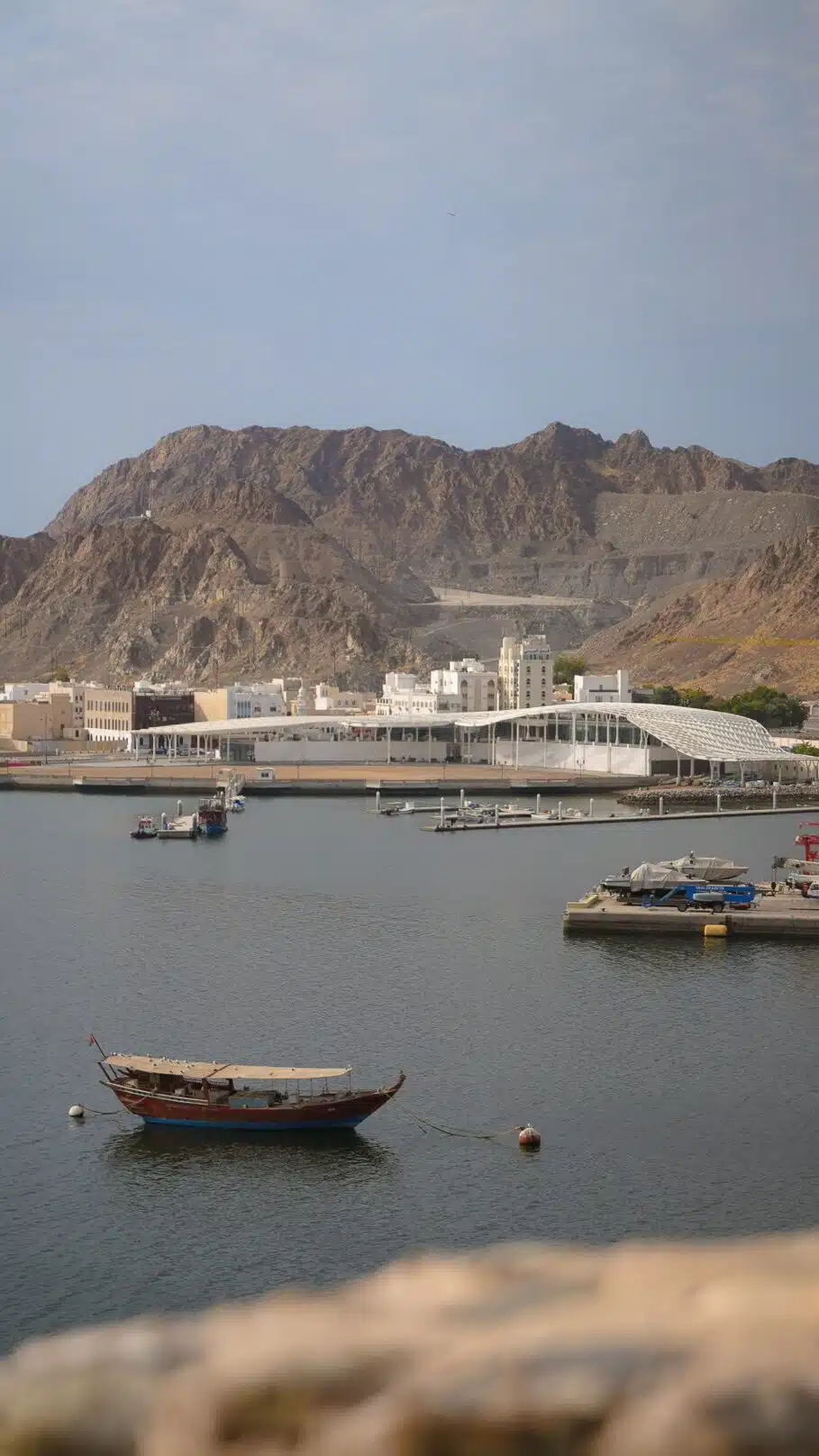 A calm harbor scene with a traditional wooden boat floating in the foreground, reminiscent of stops on a 1-week Oman itinerary. In the background, modern buildings and mountains are visible under a partly cloudy sky.