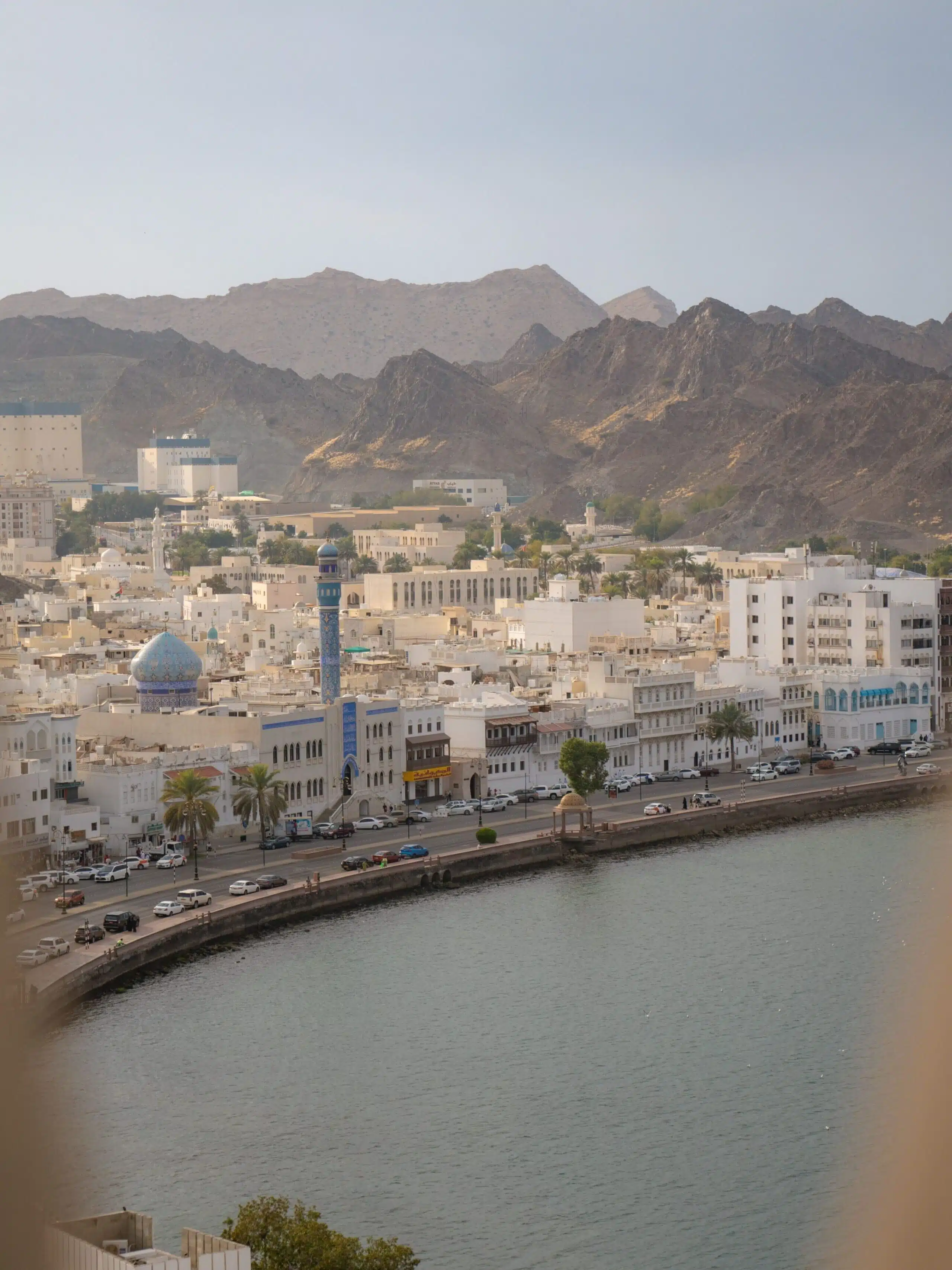 Scenic view of a city with white buildings along a waterfront, surrounded by rugged mountains—a must-see in your 1-week Oman itinerary. A blue minaret stands out among the structures, and an arch frames the serene scene under a clear sky.
