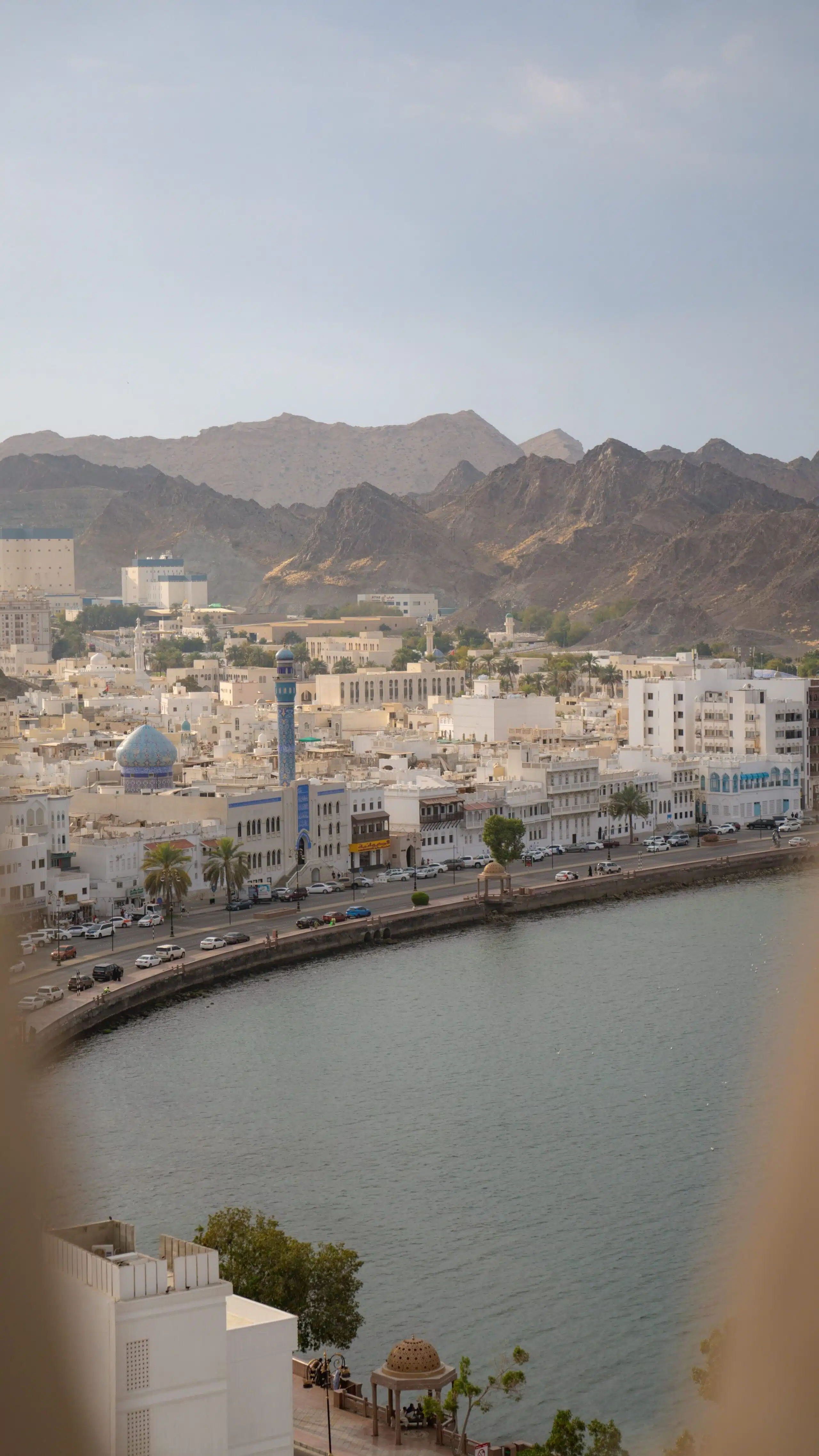 Scenic view of a city with white buildings along a waterfront, surrounded by rugged mountains—a must-see in your 1-week Oman itinerary. A blue minaret stands out among the structures, and an arch frames the serene scene under a clear sky.