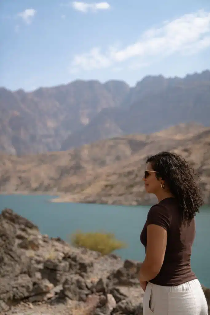 A woman with curly hair, wearing sunglasses and a brown shirt inspired by What to Wear in Oman, stands on rocky terrain overlooking a turquoise lake with mountains in the background under a partly cloudy sky.