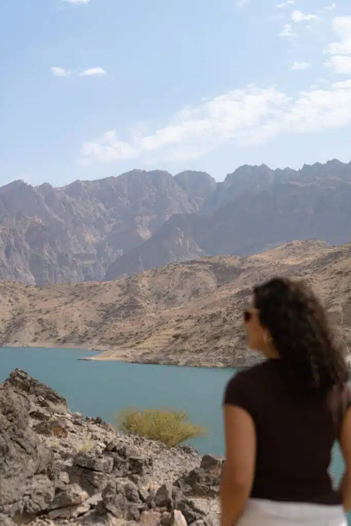 A woman with curly hair and sunglasses stands on rocky terrain, overlooking a turquoise lake surrounded by rugged mountains under a partly cloudy sky.