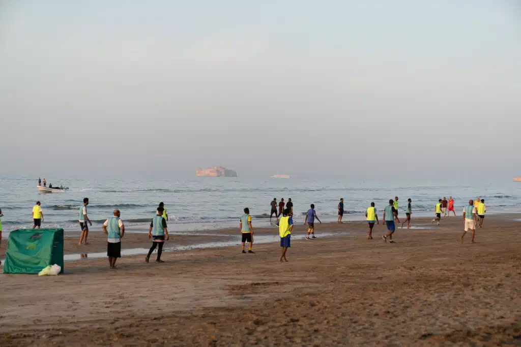 A group of people wearing sports jerseys play soccer on a sandy beach near the shoreline in Muscat, with the sea and distant islands in the background—perfect for those wondering where to stay in Muscat for seaside activities.