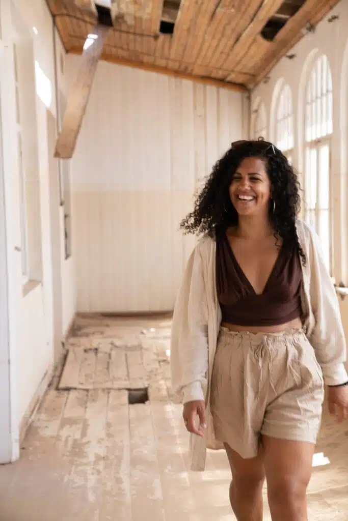 A woman with curly hair smiles while walking through a sunlit, rustic hallway with wooden floors and arched windows. She is wearing a brown top, beige shorts, and a light jacket. The ceiling and floor appear weathered.