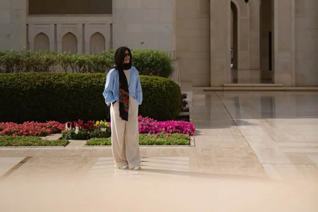 A person wearing a blue striped shirt, light pants, and a black scarf stands on a marble pathway near trimmed bushes and pink flowers outside a light-colored building with arched architectural details.
