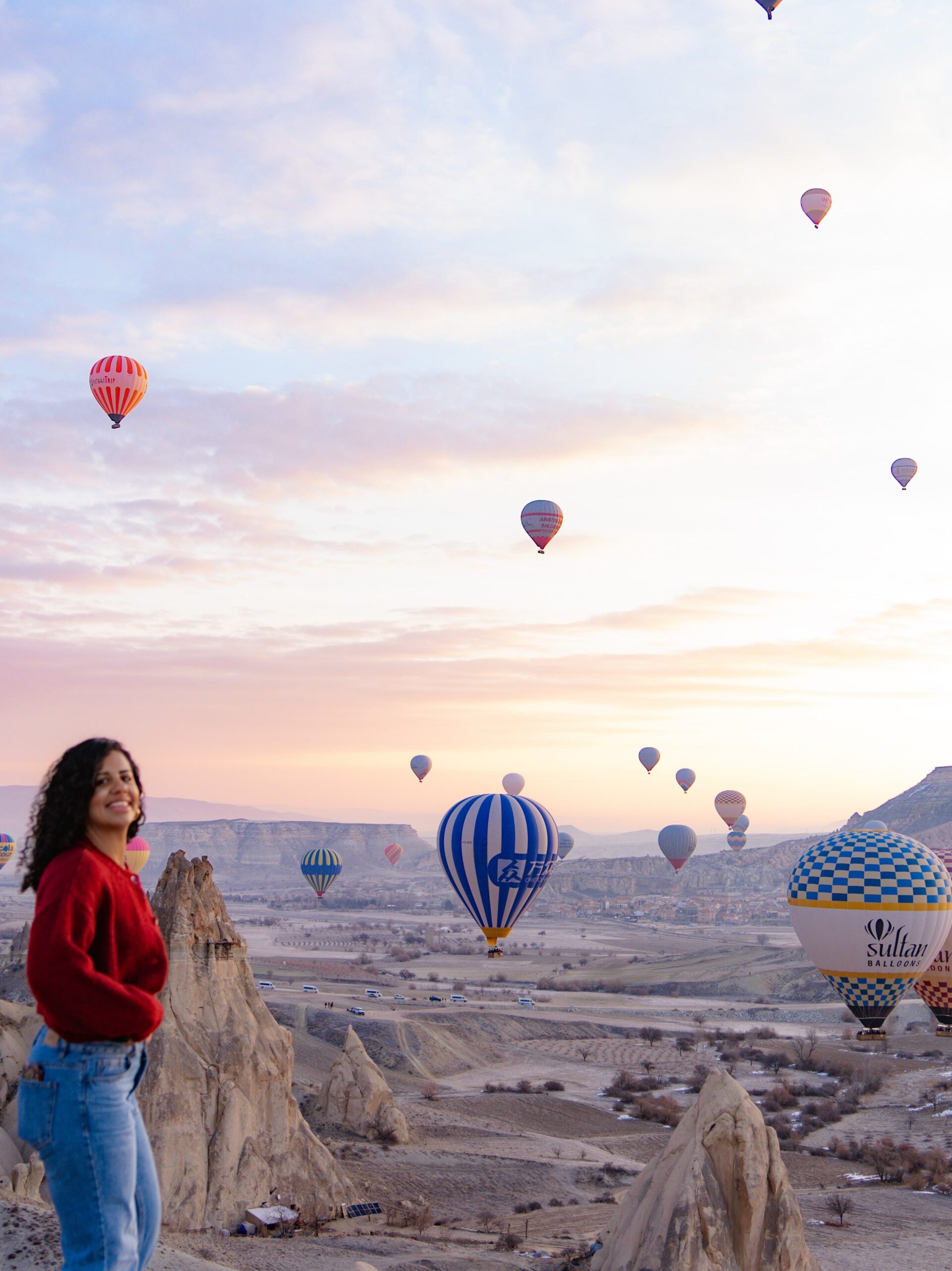 A woman in a red sweater and jeans smiles while standing on rocky terrain, with numerous colorful hot air balloons floating in the sky at sunrise or sunset.
