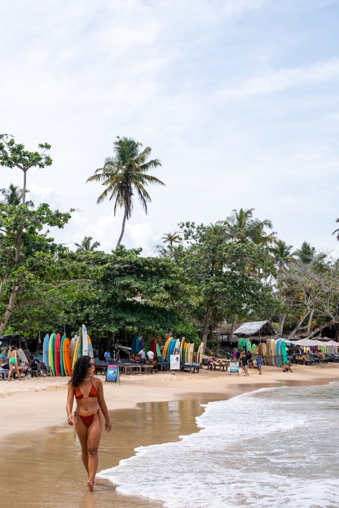 A woman in a red bikini walks along a sandy beach near the shoreline. Colorful surfboards are lined up by a beachside shop, with palm trees and people relaxing in the background under a partly cloudy sky.