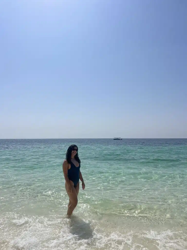 A woman in a dark swimsuit stands at the edge of clear, turquoise ocean water under a bright, cloudless sky—an inspiring scene for those wondering What to Wear in Oman’s beautiful seaside destinations. A boat drifts on the distant horizon.