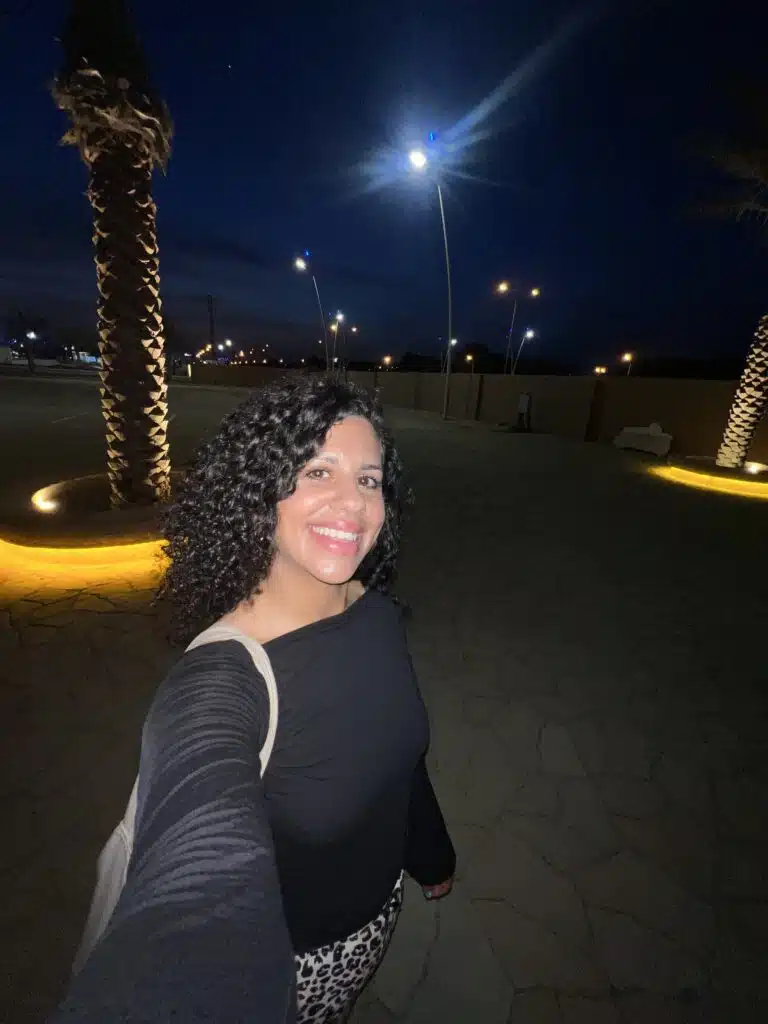 A woman with curly hair takes a selfie outdoors at night, smiling under streetlights near palm trees on a paved walkway—her outfit subtly hints at what to wear in Oman for cool, calm evening strolls.