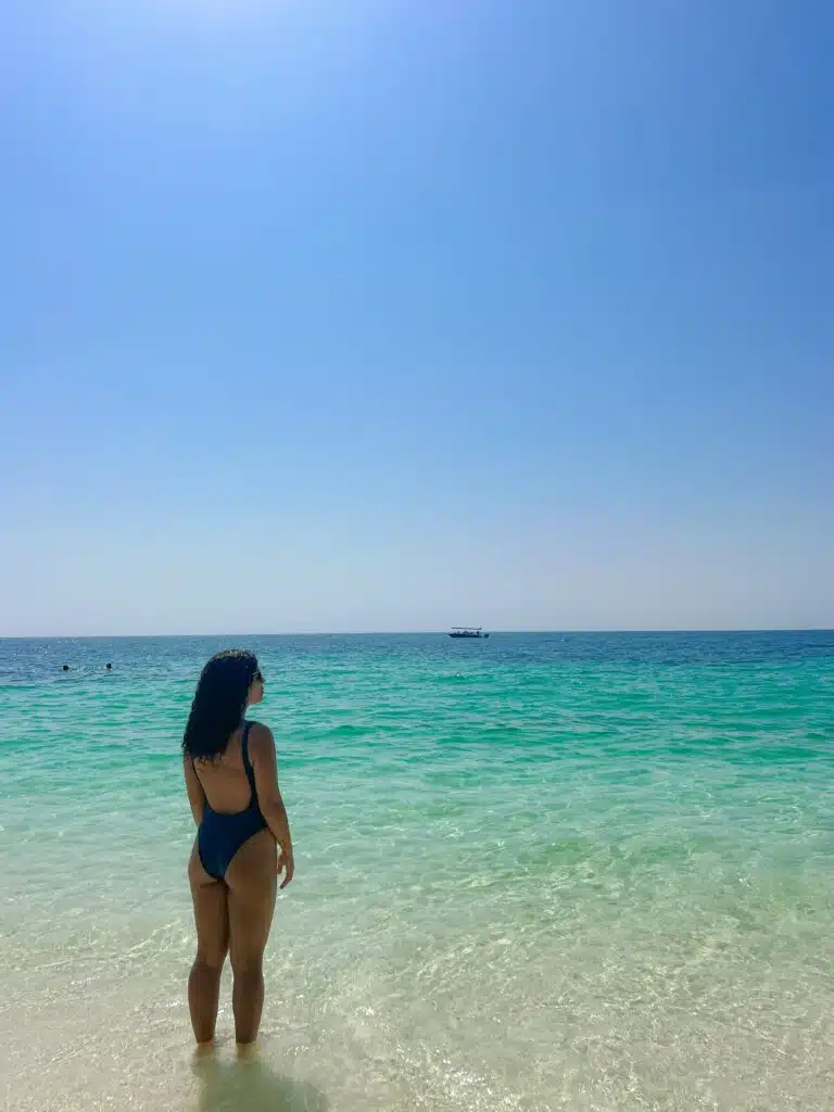 A woman in a dark swimsuit stands in shallow, clear turquoise water on a sunny beach, looking out at the calm sea. Wondering what to wear in Oman? This serene scene highlights simple, stylish beach attire under a clear blue sky. A boat drifts in the distance.