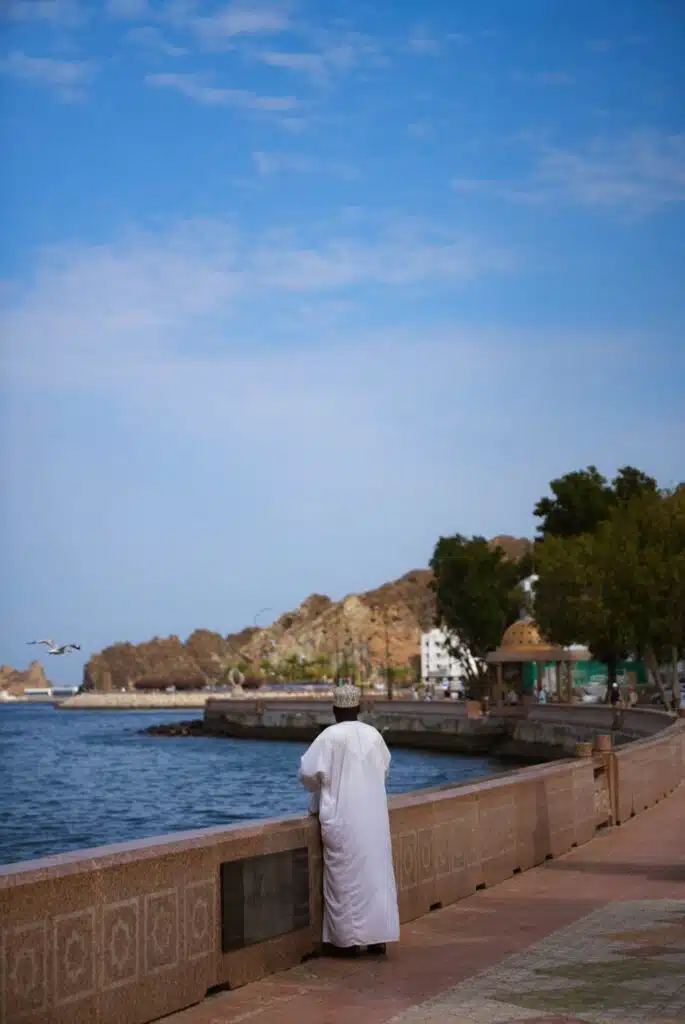 A person dressed in a white robe, typical of what to wear in Oman, stands by a seaside promenade, gazing over the water with rocky hills, trees, and buildings visible under a partly cloudy sky.
