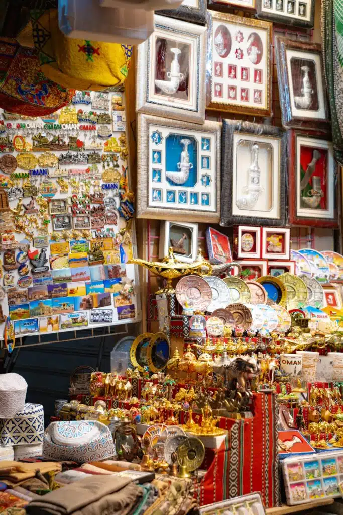 A vibrant market stall displays colorful Omani souvenirs—framed art, magnets, postcards, keychains, miniature daggers, and decorative plates—arranged on patterned fabric, perfect mementos for anyone spending 24 hours in Muscat.