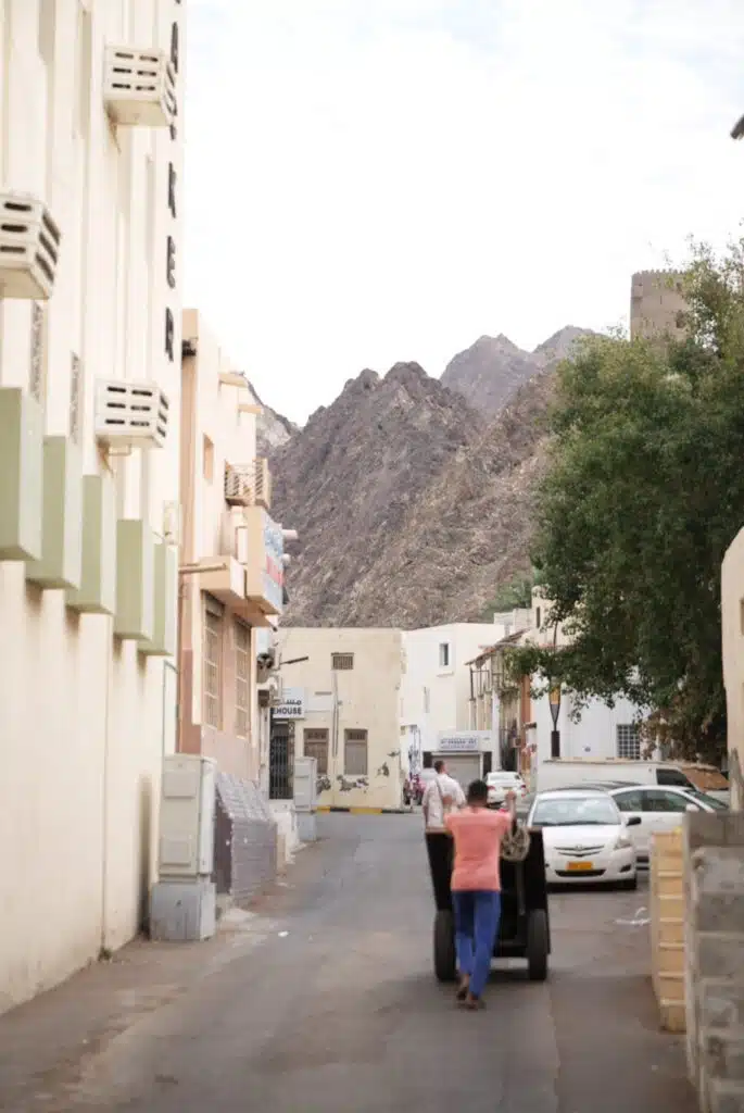 Two people push a cart down a narrow street lined with buildings, parked cars on the right, and rugged mountains in the distance—capturing a lively moment you might see during 24 hours in Muscat under a partly cloudy sky.