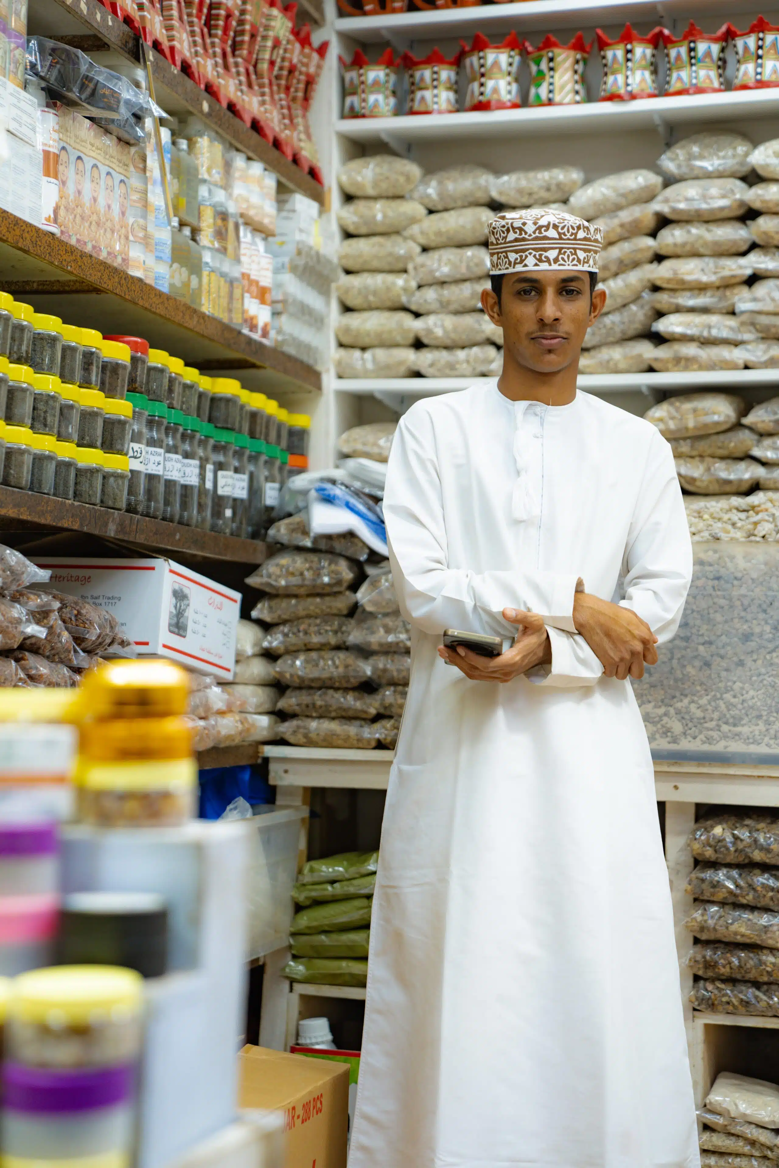 A man in traditional white clothing and cap stands in a shop filled with jars and bags of spices and herbs, holding a smartphone. Experience this vibrant scene during 24 hours in Muscat.