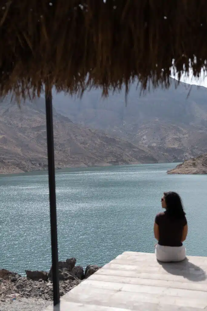 A woman with curly hair, considering what to wear in Oman, sits on a wooden platform by a calm lake, facing distant rocky mountains under a clear sky. A thatched roof partially shades the tranquil scene from above.