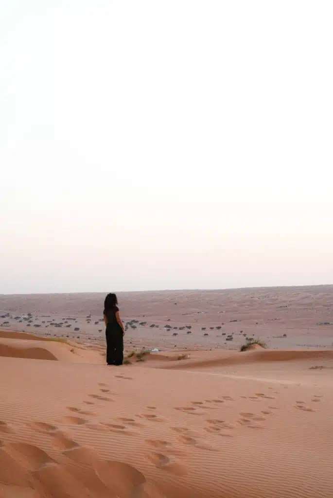 A person in dark clothing, reflecting ideas of what to wear in Oman, stands alone on sandy desert dunes at sunset. Footprints lead across rippled sand, and sparse vegetation dots the vast, empty landscape under a pale sky.