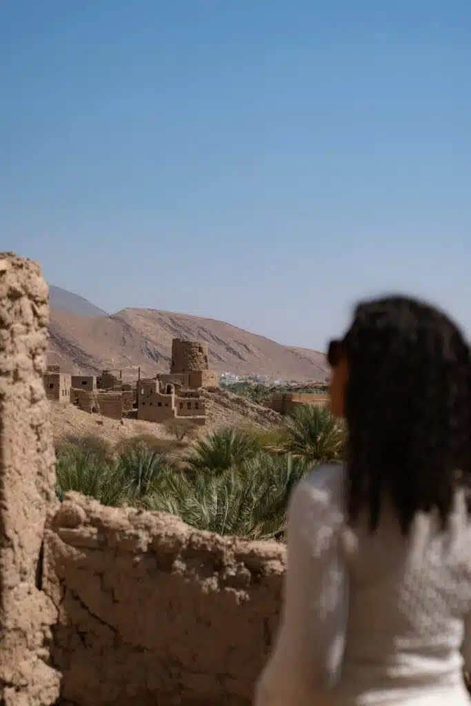 A person with curly hair and sunglasses looks out over ancient stone buildings and palm trees, considering What to Wear in Oman, with mountains and a clear blue sky in the background.
