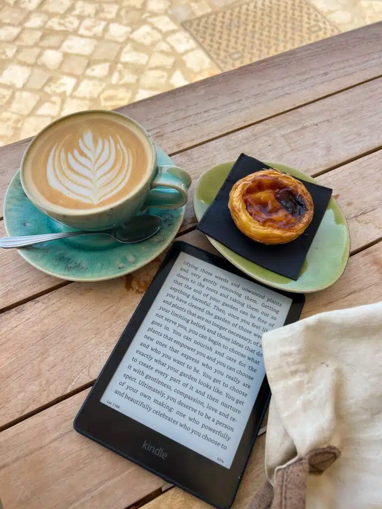 A cup of latte with latte art, a small custard tart on a green plate, and an e-reader displaying text sit atop a wooden table—perfect for a Digital Nomad in Ericeira. Part of a white bag and stone pavement are visible in the background.