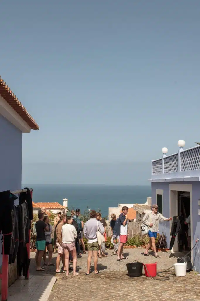 A group of people stand and talk in a sunny courtyard between pastel buildings, with the ocean visible in the background under a clear sky. Buckets and wetsuits hint at surf culture—perfect for any Digital Nomad In Ericeira seeking inspiration.
