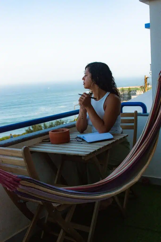 A woman, a true digital nomad in Ericeira, sits at a small wooden table on a balcony overlooking the ocean. An open notebook and cup rest nearby, with a striped hammock adding to the calm and contemplative scene.