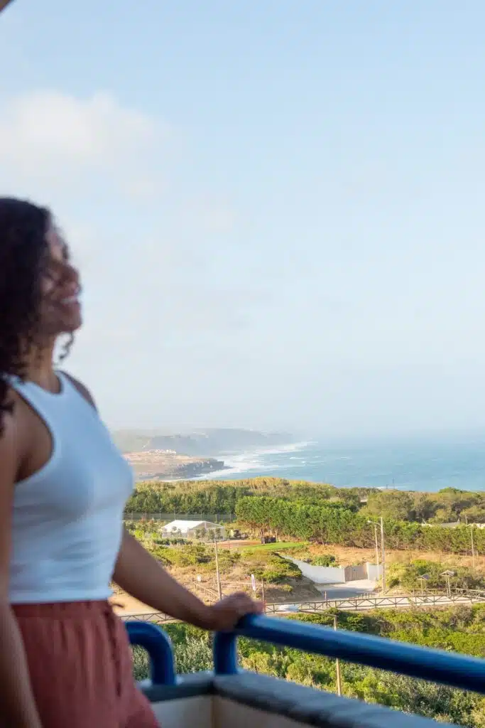 A digital nomad in Ericeira, dressed in a white sleeveless top, stands on a balcony overlooking the scenic coastline with green hills, blue ocean, and a clear sky in the background.