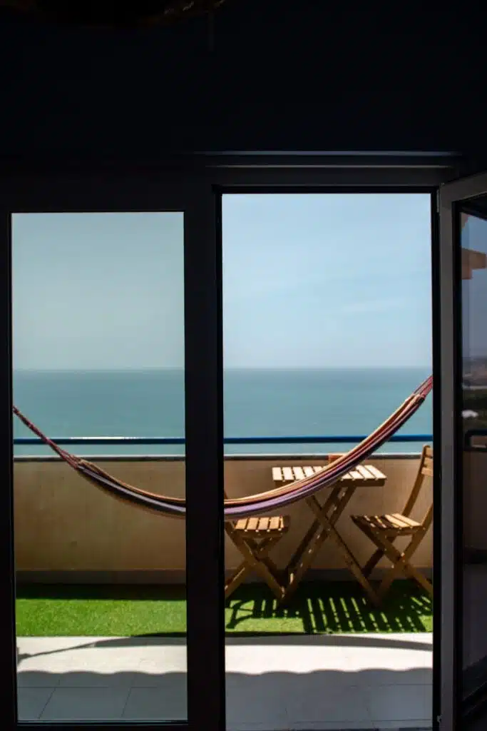 A hammock and two wooden chairs with a small table are set on a balcony overlooking a calm blue sea, viewed through glass doors—an inviting spot for any digital nomad in Ericeira.
