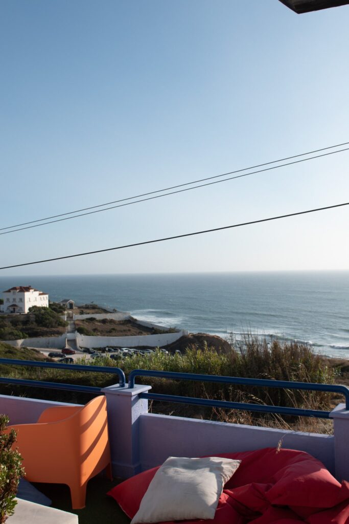 A coastal view in Ericeira with the ocean in the background, a house on the left, and a terrace in the foreground featuring an orange chair and red and white cushionsโperfect for a digital nomad in Ericeira. Power lines cross the sky above.