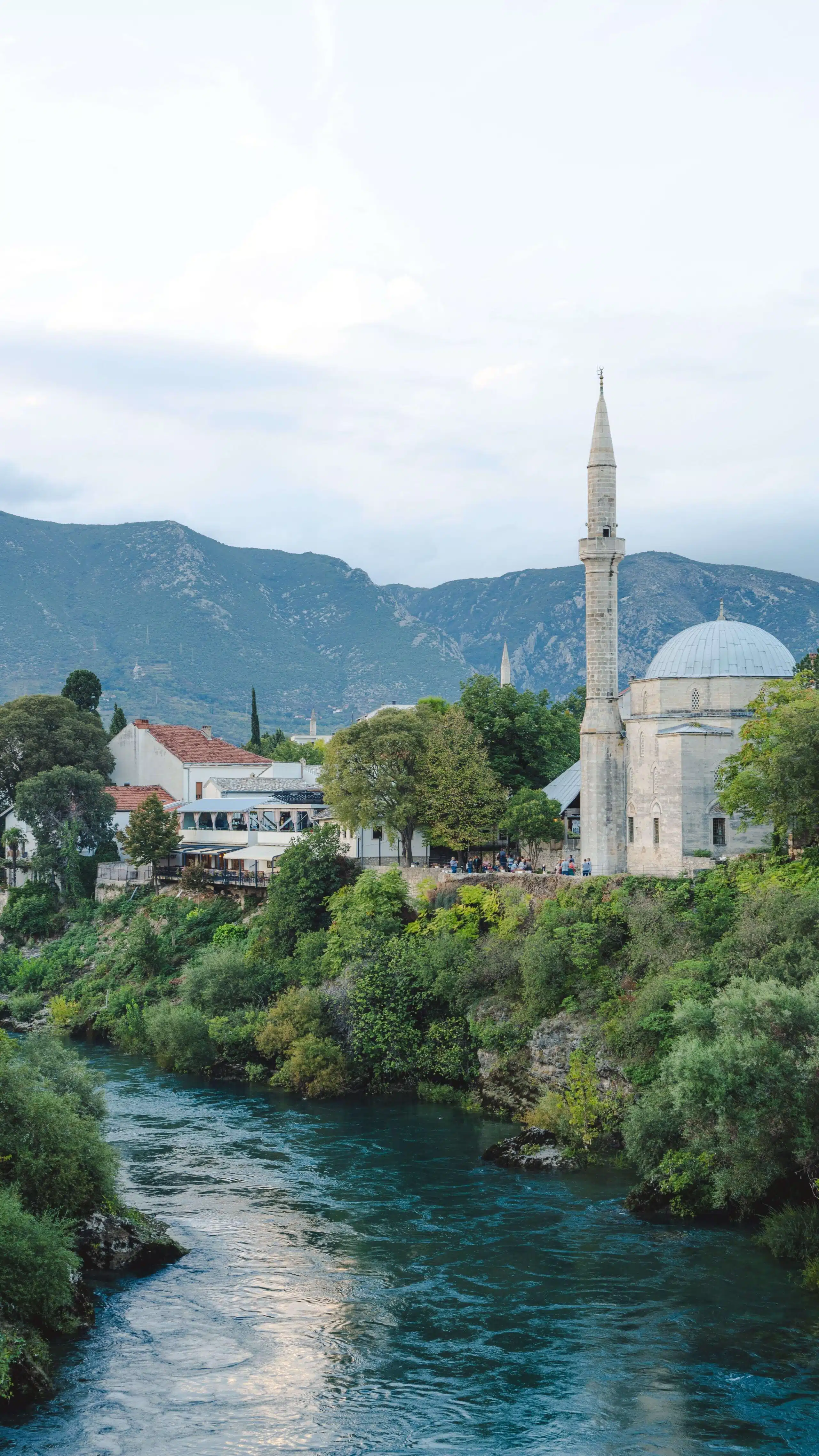 A scenic view of a river flowing through a lush landscape unfolds One Day In Mostar, with a mosque featuring a tall minaret on the riverbank. In the background, buildings and tree-covered mountains rise under a cloudy sky, painting a serene picture of this picturesque location.