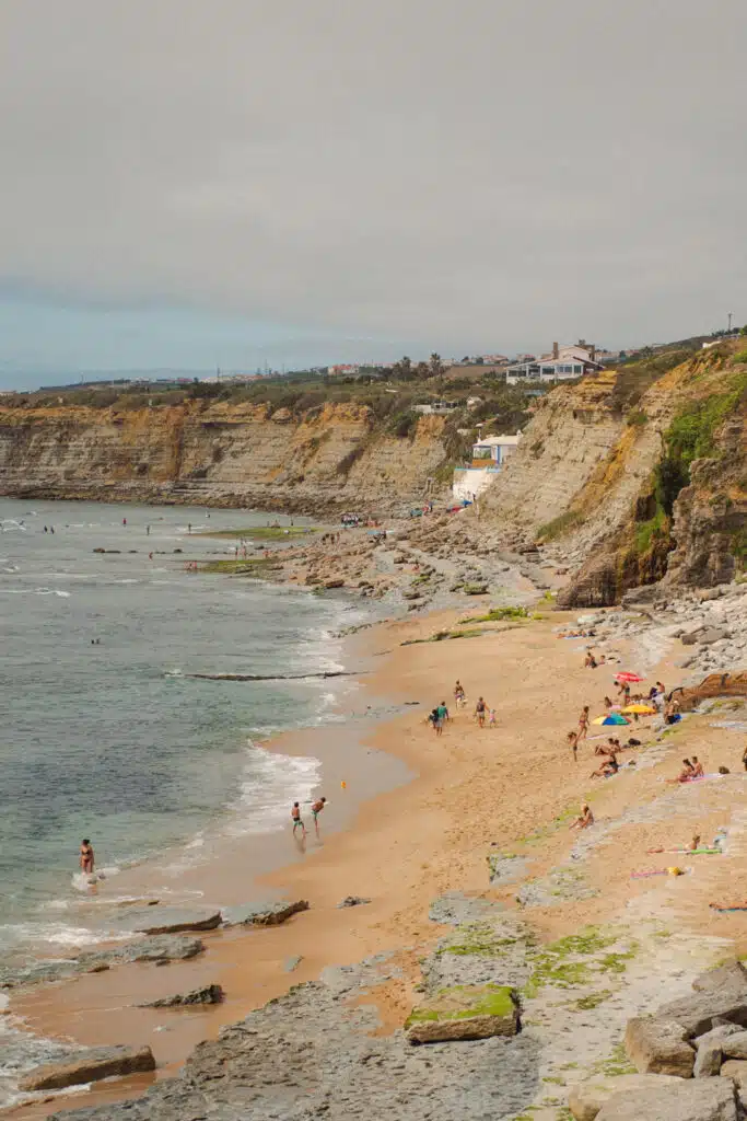 People relax and play on a sandy beach bordered by rocky cliffs and the ocean—one of the Best Beaches in Ericeira. Some swim, while others sit under umbrellas or stroll along the shore. Houses and buildings are visible atop the distant cliffs.